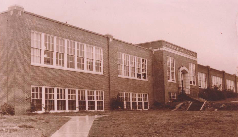 Sepia-toned photograph showing an angled view of the old brick Centreville Elementary School. The building is two stories tall and has a sidewalk and concrete stairway leading to an arched entryway. The rows of windows are painted white and two children are visible, peering out from two open windows in the distance. 