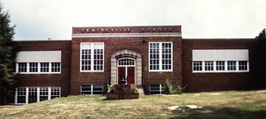 Undated color photograph of the first brick Centreville Elementary School building. The bell that used to be on the three-room schoolhouse is mounted on a brick platform in front of the main entrance.