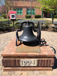 2018 color photograph of the bell at its new location in front of the modern Centreville Elementary School building. The bell is set on a raised brick platform and with a bronze plaque inscribed with its history. The bell can be rung by turning the wheel attached to its right side. 